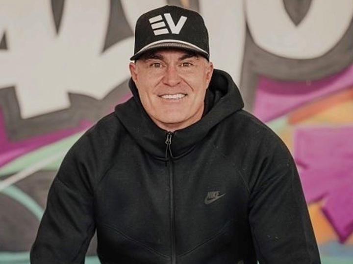 Photo of EV Auto owner Alex Lawrence wearing a black ballcap and black hoodie sweatshirt in front of a graffiti mural
